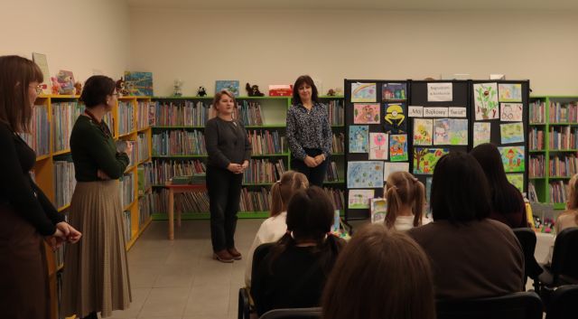 na zdjęciu dyrekotrka biblioteki i komisja kponkurowa stoją przed siedzącymi na krzesłach dziećmi z rodzicami i nauczycielami, w tle wystawa nagrodzonych prac konkursowych i regały z książkami