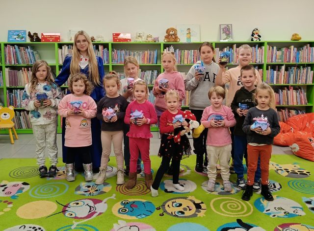 dzieci i bibliotekarka w stroju księżniczki pozują do zdjęcia ze swoimi słoikami z miasteczkami smerfów, w tle regały z książkami