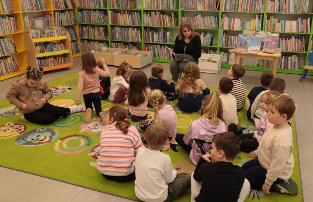 dzieci siedzą na zielonym dywanie, przed nimi na krzesełku siedzi bibliotekarka z książką, w tle regały z książkami