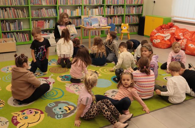 dzieci siedzą na zielonym dywanie, przed nimi na krzesełku siedzi bibliotekarka z książką, w tle regały z książkami
