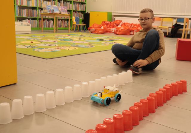 chłopiec siedzi po turecku na podłodze, przed nimbiałe i czerwone kubeczki ustawione w rzędach, między nimi robot z klocków Lego