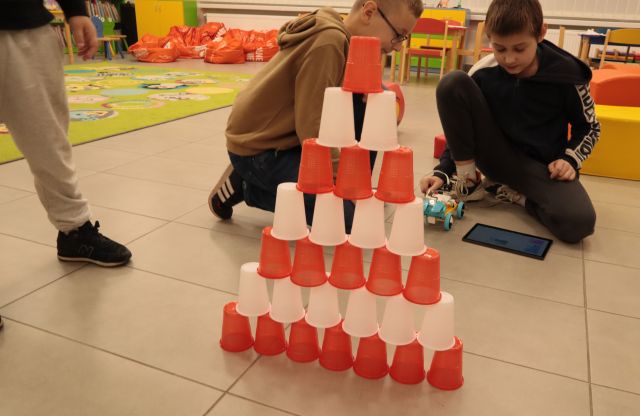 na podłodze piramida z białych i czerwonych kubeczków, za nimi na podłodze siedzą dzieci