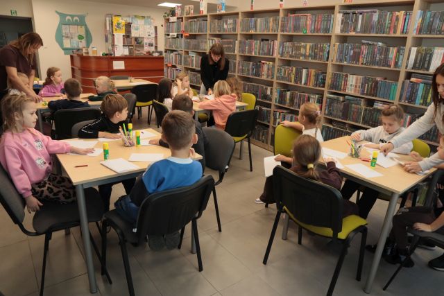 przedszkolaki siedzą przy stołach, na stołach leżą kartki, w tle lada biblioteczna i regały z książkami