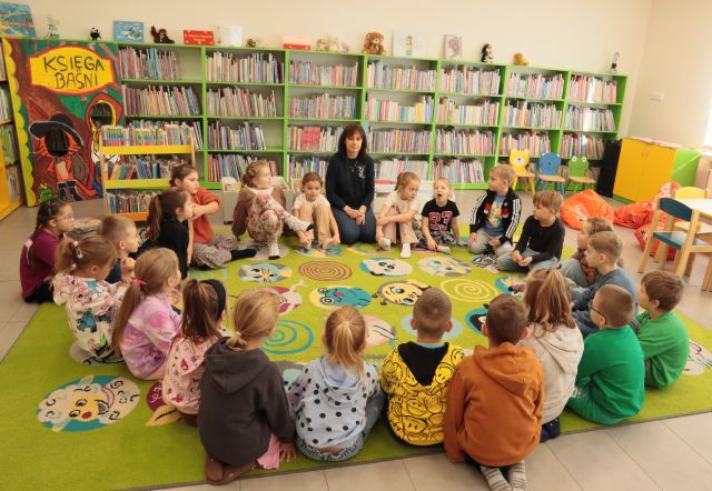 przedszkolaki i dyrektorka biblioteki siedzą na zielonym dywanie, w tle regały z książkami