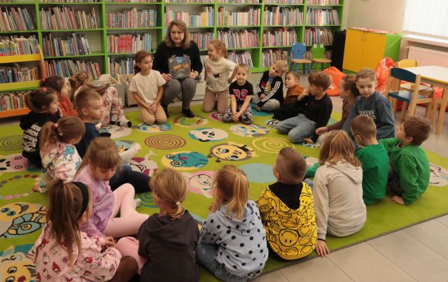 przedszkolaki siedzą na zielonym dywanie, między nimi na krzesełku siedzi bibliotekarka z książką, w tle regały z książkami