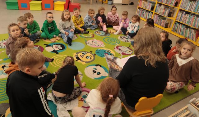 dzieci siedzą na zielonym dywanie, między nimi na krzesełku siedzi bibliotekarka z książką