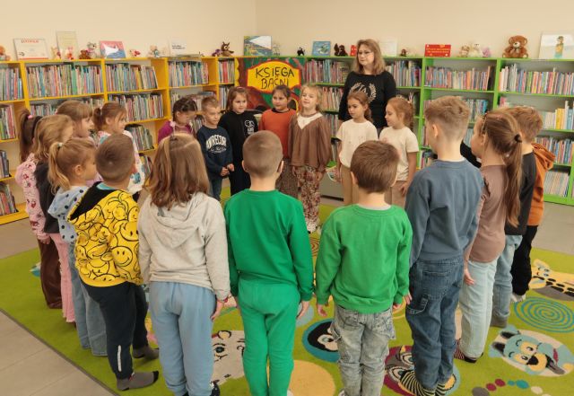przedszkolaki i bibliotekarka stoją na zielonym dywanie, w tle regały z książkami