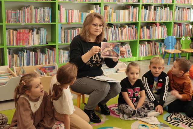 bibliotekarka siedząca na krzesełku pokazuje dzieciom zdjęcie z bajki, w tle regały z książkami