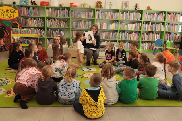 bibliotekarka siedząca na krzesełku pokazuje dzieciom siedzącym na zielonym dywanie książkę, zdjęcie misia, w tle regały z książkami
