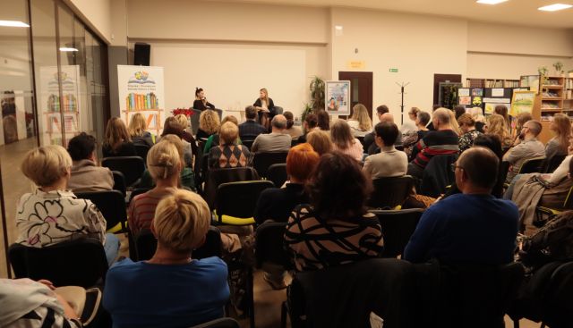 uczestnicy spotkania siedzą na krzesłach, w tle przy stole siedzi autorka z moderatorką