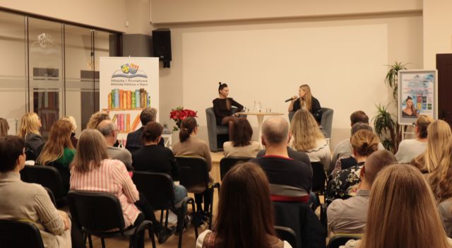 uczestnicy spotkania siedzą na krzesłach, w tle przy stole siedzi autorka z moderatorką