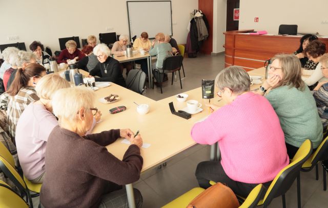 około 20 seniorek siedzi przy ustawionych w literę u stołach, rozwiązują zadania na kartkach