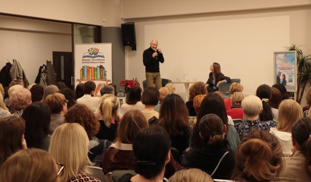 dr stoi na scenie z mokrofonem, dziennikarka siedzi na fotelu przed publicznością