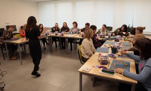 bibliotekarki siedzą przy stołach ustawionych w podkowę, na stołach kartki, słoiki, kleje, nożyczki, przed stołami stoi prowadząca szkolenie