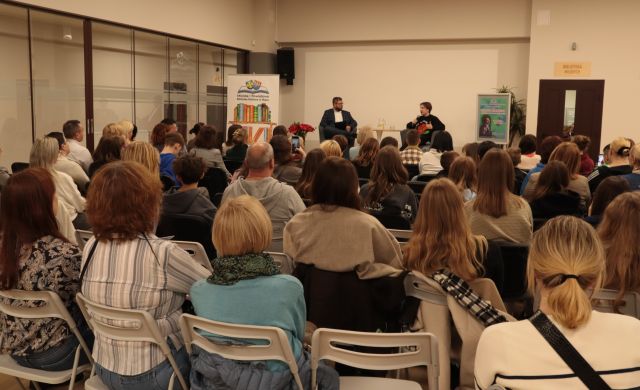 na zdjęciu publiczność siedząca na krzesłach, w tle stoją: dyrektorka biblioteki, prowadzący spotkanie i gość spotkania