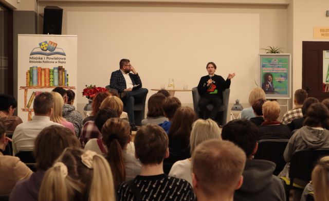 na zdjęciu publiczność siedząca na krzesłach, w tle stoją: dyrektorka biblioteki, prowadzący spotkanie i gość spotkania