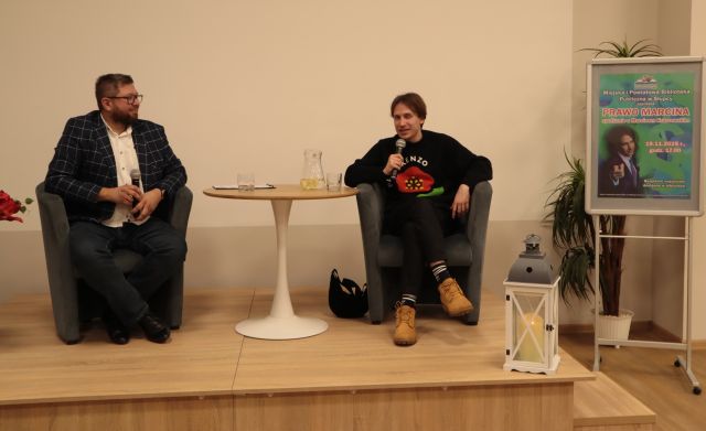 na scenie na fotelach przy okrągłym stoliku siedzi prowadzący spotkanie i gość