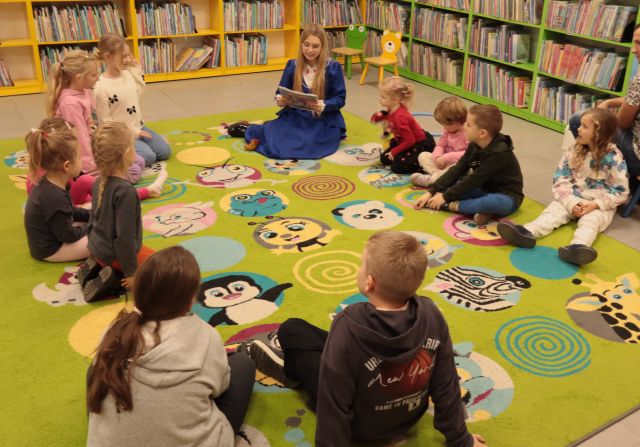 dzieci i bibliotekarka w sukience księżniczki i książką siedzą na kolorowym dywanie