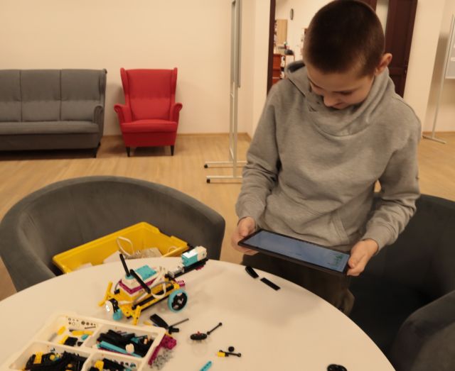 chłopiec tabletem, przed nim na stoliku pojazd z klocków LEGO