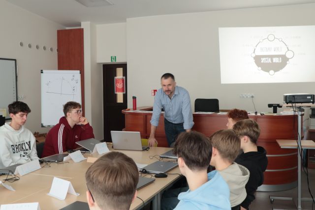 szkoleniowiec oparty o stół mówi do młodzieży siedzącej przy komputerach