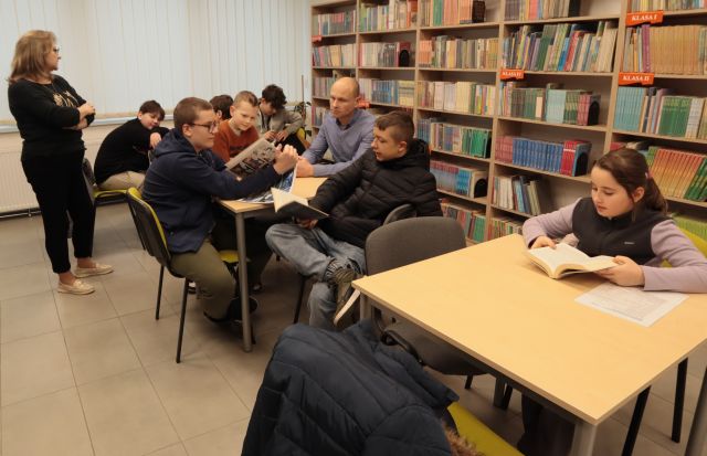 młodzież z nauczycielem siedzą przy stołach w sali bibliotecznej