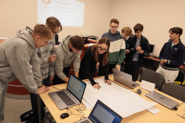 młodzież pochylona nad dużym arkuszem papieru, dziewczyna rysuje na nim, na stołach laptopy