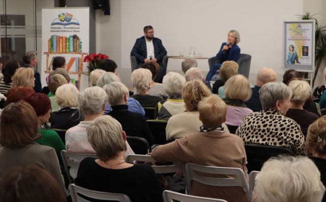 na zdjęciu uczestnicy spotkania siedzący na krzesłach, w tle siedzący na fotelach mężczyzna i kobieta