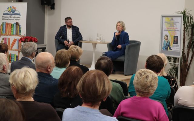 na zdjęciu uczestnicy spotkania siedzący na krzesłach, w tle siedzący na fotelach mężczyzna i kobieta