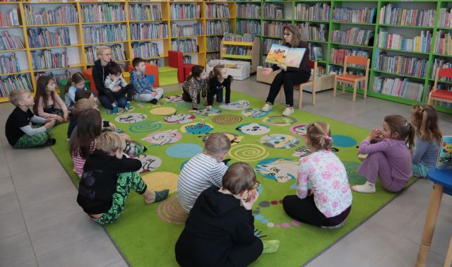 przed dziećmi siedzącymi na kolorowym dywanie na krzesełku siedzi bibliotekarka z książką, w tle regały z książkami
