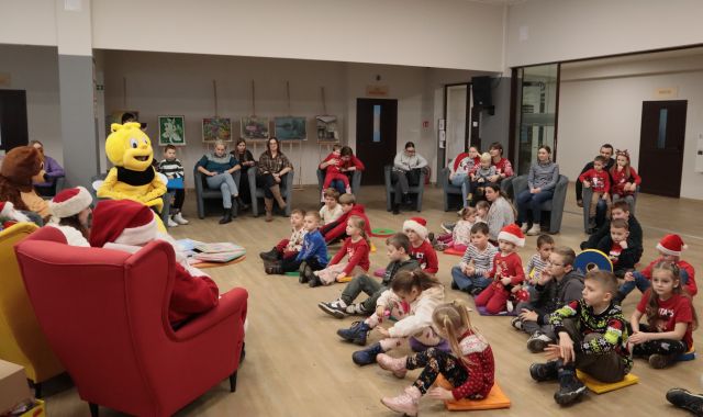 dzieci siedzą na podłodze na pufach, na fotelach siedzą rodzice, przed dziećmi siedzą w fotelach Mikołaj, pani Mikołajowa, Miś i Pszczółka, w tle duża choinka