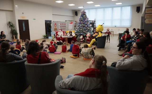 dzieci siedzą na podłodze na pufach, na fotelach siedzą rodzice, przed dziećmi siedzą w fotelach Mikołaj, pani Mikołajowa, Miś i Pszczółka, w tle duża choinka
