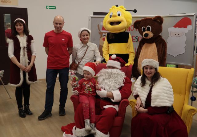 małe dziecko siedzi na kolanach Mikołaja, obok siedzi Mikołajowa, za nimi stoi pomocnica Mikołaja, mężczyzna w czerwonej bluzce, kobieta w czapce Mikołaja, Pszczółka i Miś