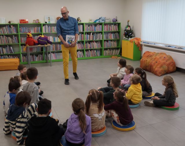 Na zdjęciu znajduje się mężczyzna z książką w ręku. Przed nim w półokręgu siedzi grupka przedszkolaków. W tle znajdują się półki z książkami