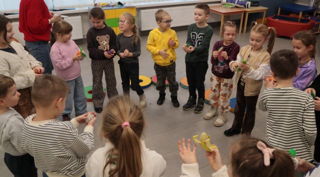 Na zdjęciu znajduje się grupa przedszkolaków stojąca w kole, ręce mają wyciągnięte przed siebie i trzymają w nich po jednym małym, zabawkowym owocu lub warzywie. Za nimi na podłodze leżą okrągłe pufy oraz stoi komoda..