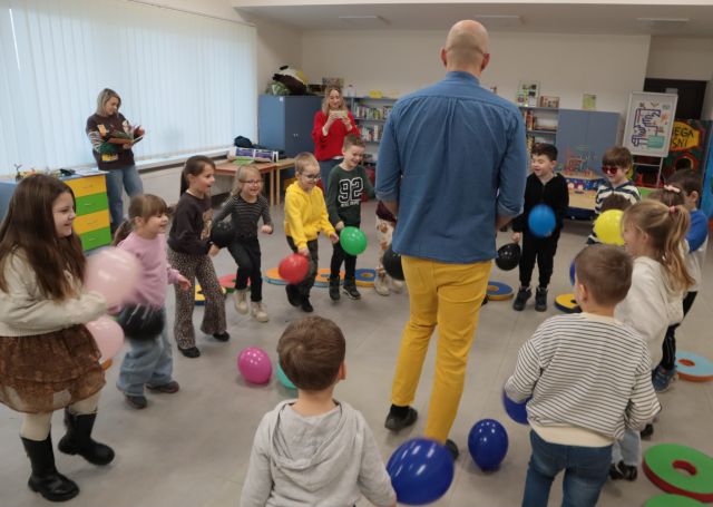 Na zdjęciu znajdują się przedszkolaki w okręgu, są w ruchu i trzymają w ręce kolorowe balony. Poza kołem stoi kobieta w czerwonym swetrze oraz kobieta stojąca przy oknie, przeglądająca książkę. W okręgu stoi mężczyzna. W tle znajdują się komody, półki z grami.