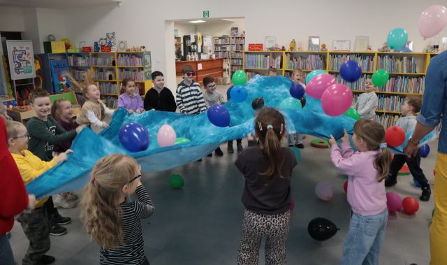 Na zdjęciu znajdują się przedszkolaki, które ruszają niebieską chustą, na której znajdują się kolorowe balony. W tle znajdują się półki z książkami.