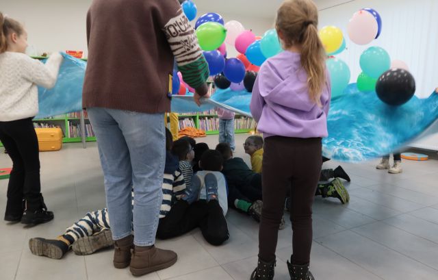 Na zdjęciu znajdują się dzieci oraz jedna kobieta, które trzymają niebieską chustę. Nad chustą unoszą się kolorowe balony, a pod chustą leżą chłopcy. W tle znajdują się półki z książkami oraz okna.