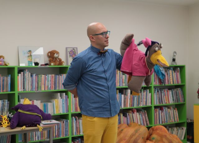 Na zdjęciu znajduje się mężczyzna trzymający przed sobą, na ręce maskotkę kaczki w różowej sukience. W tle znajdują się półki z książkami, oraz stolik.