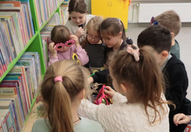 Na zdjęciu znajduje się ośmioro przedszkolaków wybierających i przymierzających różne okulary ze stolika, który znajduje się przy półce z książkami. W tle znajduje się żółta szafka i kaloryfer.