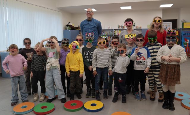 Na zdjęciu znajduje się grupa 17 przedszkolaków, za nimi na środku stoi mężczyzna w niebieskiej koszuli, a bliżej prawej strony znajdują się dwie kobiety. Wszyscy mają na sobie kolorowe okulary. Przed nimi leżą kolorowe, okrągłe pufy, za nimi znajdują się okna oraz półki z grami.