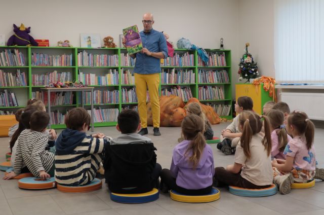 Na zdjęciu znajduję się mężczyzna stojący z otwartą książką. Przed nim w półokręgu siedzą przedszkolaki. w tle znajdują się półki z książkami.