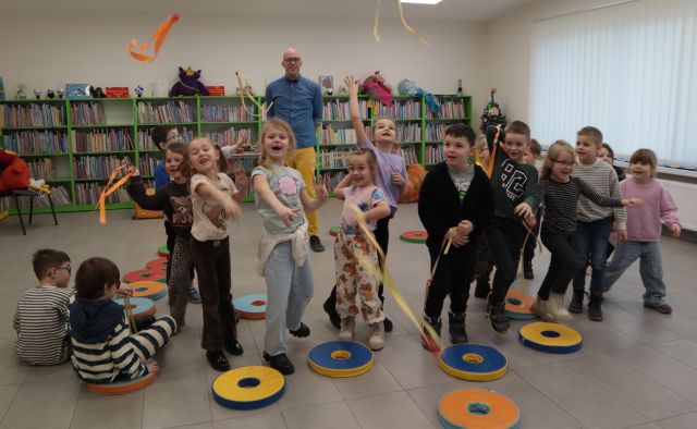 Na zdjęciu znajdują się przedszkolaki, rzucające wstążkami. Po lewej stronie siedzi dwójka chłopców. Za przedszkolakami stoi mężczyzna. W tle widać półki z książkami.