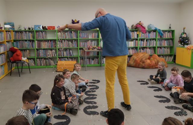 Na zdjęciu znajdują się przedszkolaki siedzące w półokręgu. Przed nimi, na podłodze znajdują się czarne skrzydła. Na środku znajduję się mężczyzna, które sypie na dzieci z góry, kolorowe konfetti. W tle znajdują się półki z książkami.