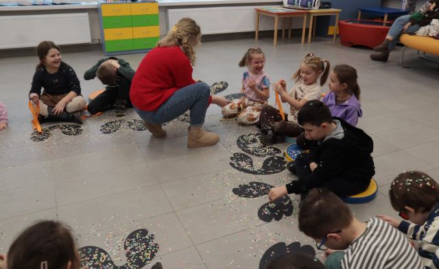 Na zdjęciu, w półokręgu siedzą przedszkolaki, które mają przed sobą czarne, papierowe skrzydła posypane kolorowym konfetti. Na środku, przy jednym dziecku kuca kobieta, w czerwonym swetrze i trzyma skrzydła w ręce. W tle znajduję się szafka oraz dwa złączone stoliki.