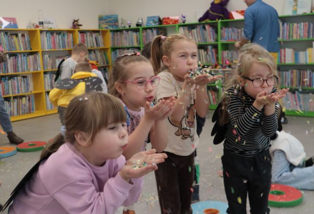 Na zdjęciu widoczne są cztery dziewczynki, lekko pochylone, które zdmuchują z rąk kolorowe konfetti. Za nimi widać chodzące przedszkolaki, mężczyznę stojącego przy półce z książkami oraz półki z książkami.