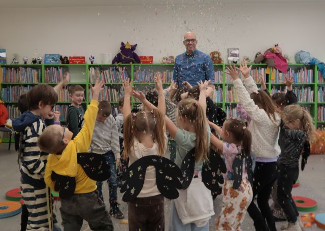Na zdjęciu znajduje się grupa przedszkolaków stojąca tyłem, na plecach mają czarne skrzydła z kolorowymi kropkami. Ręce mają podniesione do góry i rzucają kolorowym konfetti. Za nimi stoi mężczyzna, w niebieskiej koszuli. W tle znajdują się półki z książkami.