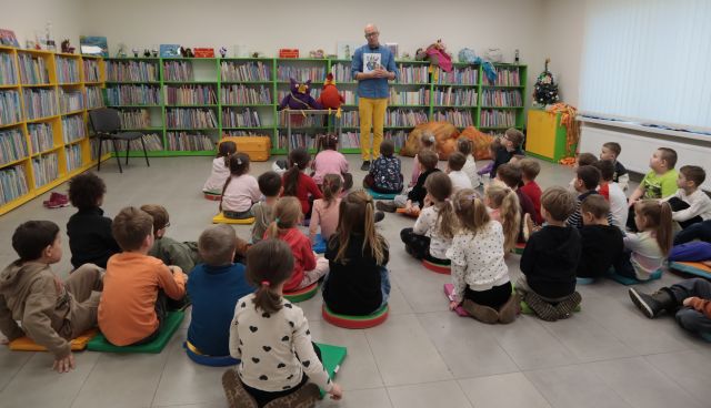Na zdjęciu znajduje się mężczyzna, trzymający w rękach książkę. Przed nim siedzą przedszkolaki. W tle znajdują się półki z książkami.