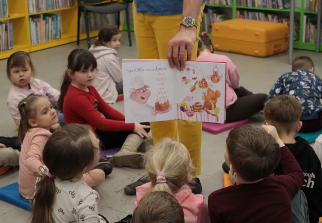 Na zdjęciu znajduje się mężczyzna od pasa w dół, trzymający w ręce otwartą książkę i pokazuje ilustrację dzieciom znajdującym się dookoła niego. W tle znajdują się półki z książkami oraz żółta walizka. 