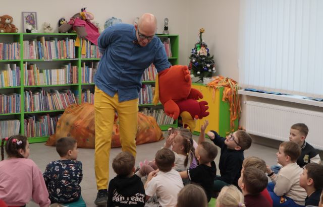 Na zdjęciu znajduję się mężczyzna trzymający na ręce pluszową maskotkę kury i pochylający się z nią w kierunku siedzących na podłodze dzieci. Za mężczyzną znajdują się półki z książkami, a po prawej stronie, z tyłu widoczna jest szafka oraz okna, a pod nimi grzejniki.