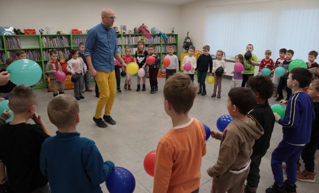 Na zdjęciu znajdują się przedszkolaki stojące w kółku, trzymające przed sobą balony. W kole znajduje się lekko podskakujący mężczyzna, również trzymający w ręce balon. W tle znajdują się półki z książkami. 